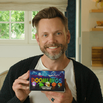 A picture of Joel McHale smiling with a Doomlings box